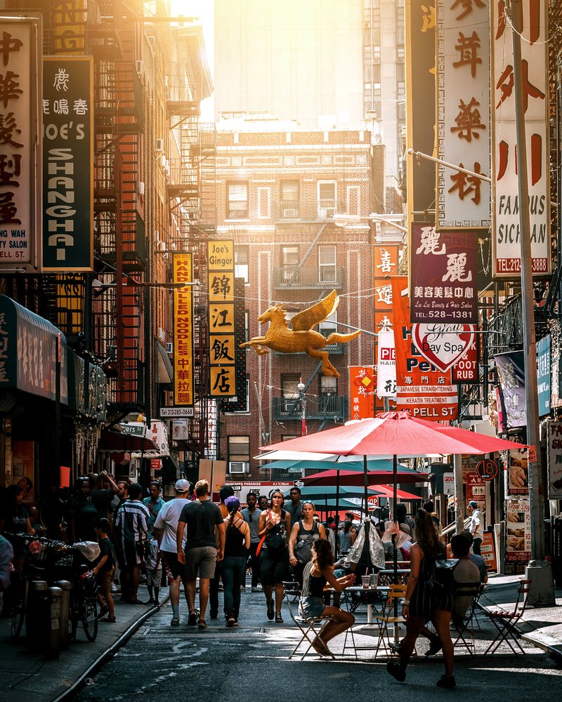 A busy city street scene with many pedestrians walking and sitting under umbrellas, with storefronts and buildings adorned with colorful signs in Chinese characters.