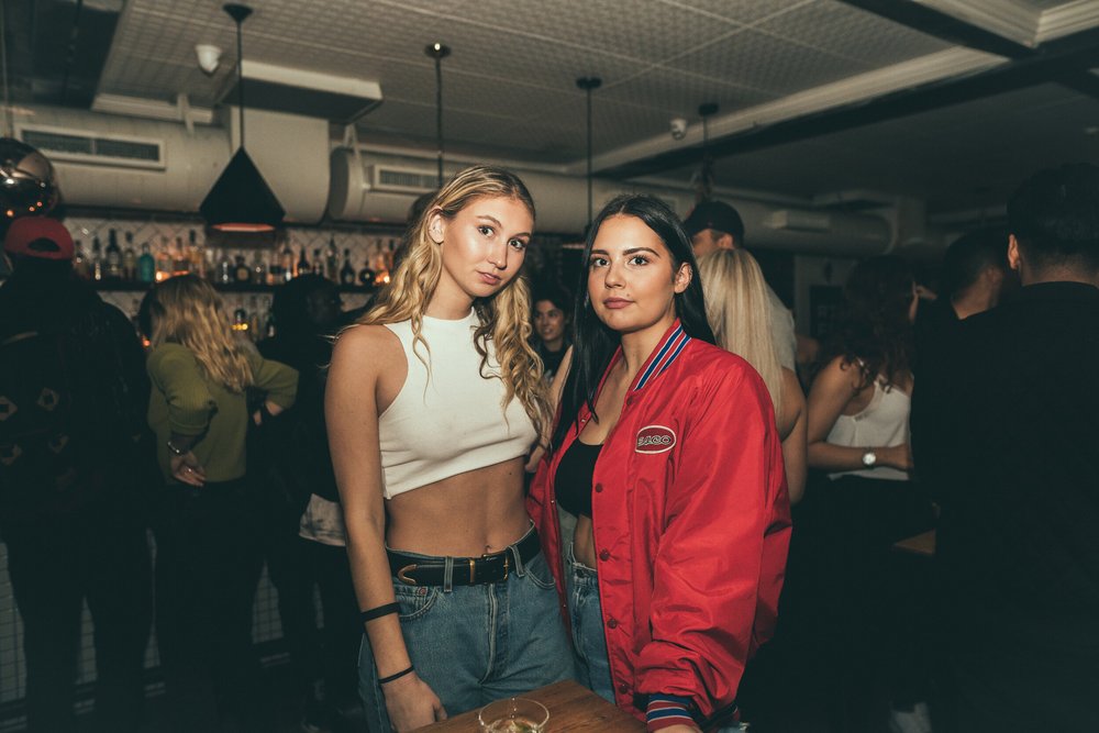 Two young women standing close together in a busy bar, one wearing a white crop top and jeans, the other dressed in a red jacket with a logo, surrounded by people socializing in the background.