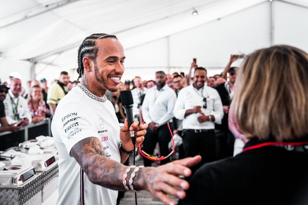 Formula 1 driver Lewis Hamilton smiling while speaking into a microphone and interacting with a woman, at an event with a crowd in the background.