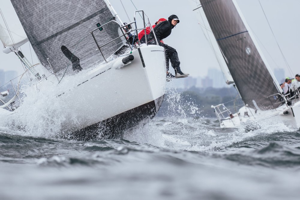 Sailboats on the water during a race, with one boat in the foreground showing sailors leaning out over the side to balance the boat in rough waters, under a gray sky.
