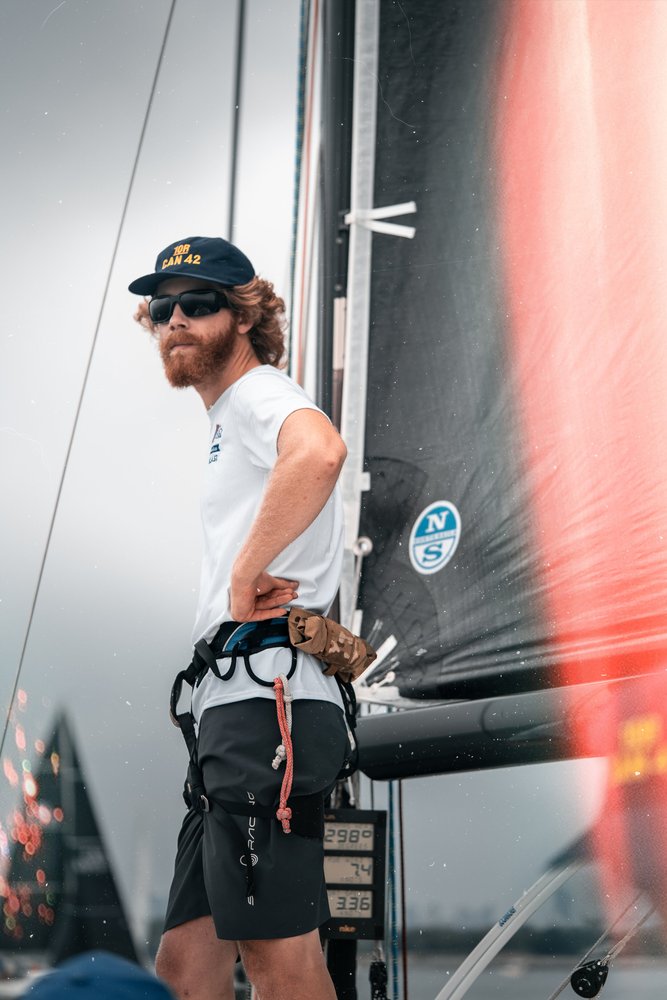 A man with a beard and curly hair wearing a black cap, sunglasses, a white shirt, and black shorts, standing next to a sailboat at a marina or dock, with a digital weather station showing temperature and other data nearby.