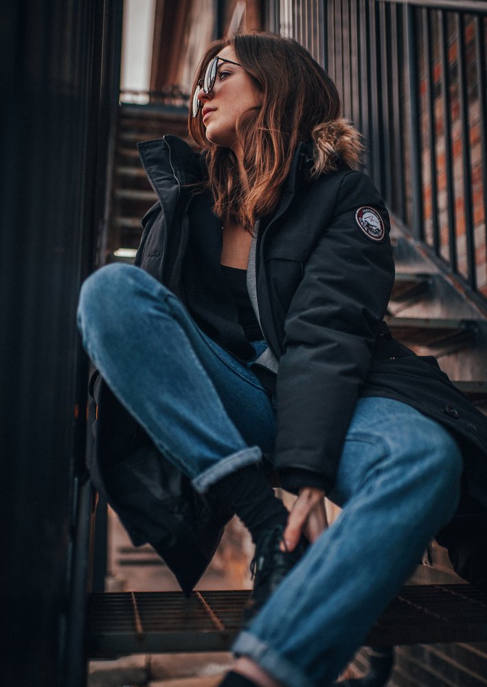 A woman sitting on a metal staircase, wearing sunglasses, a black jacket, and blue jeans, looking to the side.